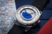 Новая модель швейцарского часового бренда De Bethune с функцией GMT создана с 42-мм корпусом из полированного титана на фирменных подвижных ушках, позволяющих часам эргономично садиться на запястье любого размера. В центре циферблата – пластина, разделенная на две части, которые символизируют день (с полированным золотым Солнцем на гильошированном небосклоне) и ночь (с Млечным Путем из десятков золотых «звезд»). Центральные стрелки из вороненой стали показывают местное время, тогда как «домашнее» время отсчитывает двухцветная сфера, двигающаяся в выемке с 24-часовой шкалой, опоясывающей центральную часть циферблата. В 6 часов утра и в 18 часов вечера сфера поворачивается, меняя цвет с синего на золотой и обратно, таким образом как бы символизируя восход и закат Солнца. Третья центральная стрелка с белым наконечником отсчитывает дату по круговой шкале. Механизм часов укомплектован титановым балансовым колесом с золотыми инерционными грузами, металлической балансовой спиралью с плоской концевой и анкерным колесом из кремния. Энергии двух заводных барабанов механизма хватает на 5 дней автономной работы.